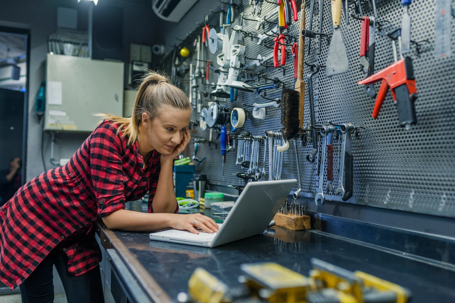 Eine Frau im rot-schwarzen Karomuster, die sich an einem Werkbanktisch in einer Werkstatt beugt. Sie arbeitet konzentriert an einem Laptop, während im Hintergrund eine Wand voller Werkzeuge zu sehen ist.