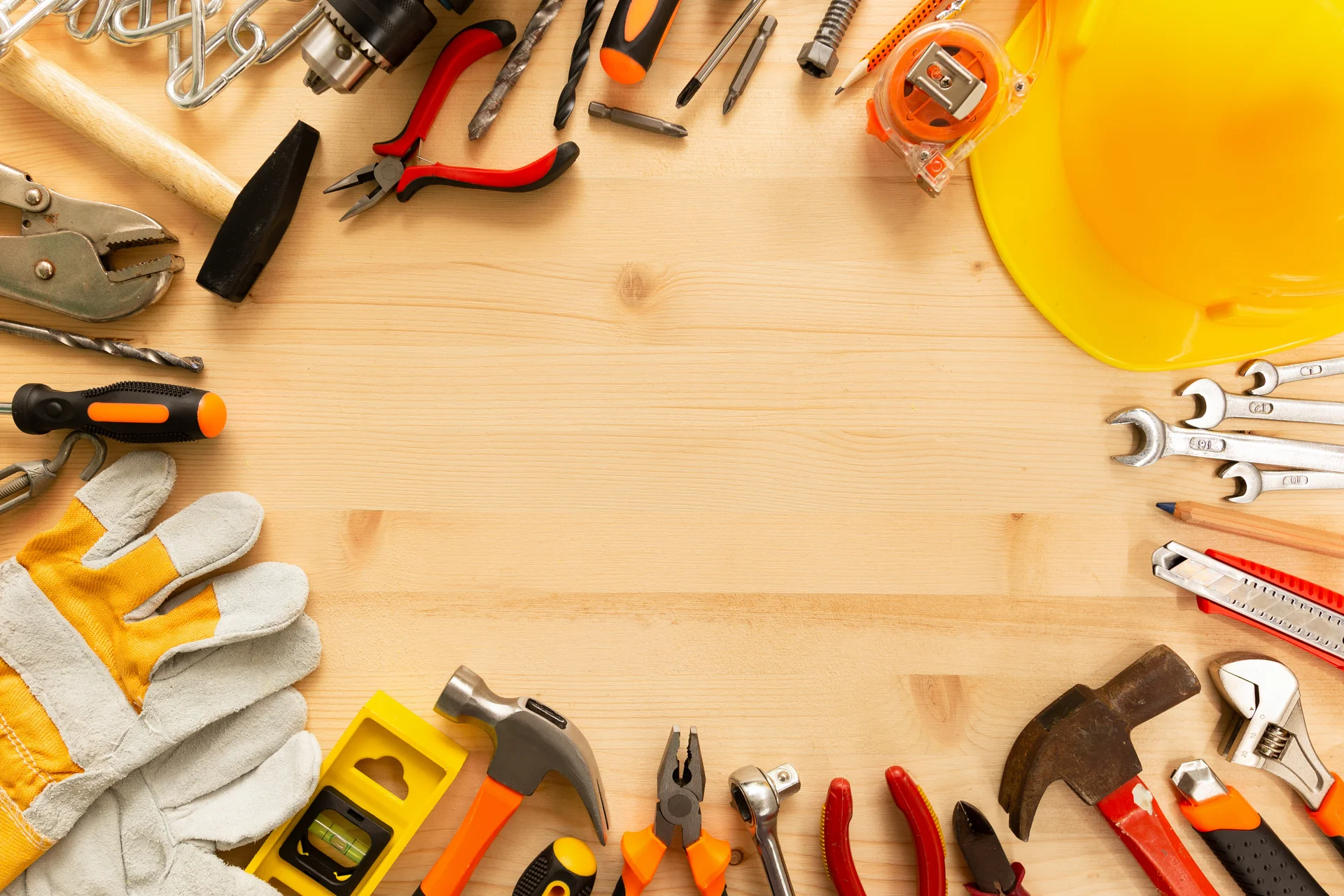 Ein flacher Blick auf einen Holztisch, der mit verschiedenen Handwerkzeugen umrandet ist. Zu sehen sind unter anderem ein Hammer, Schraubenschlüssel, Zangen und ein Bauhelm – typische Werkzeuge eines Handwerkers.