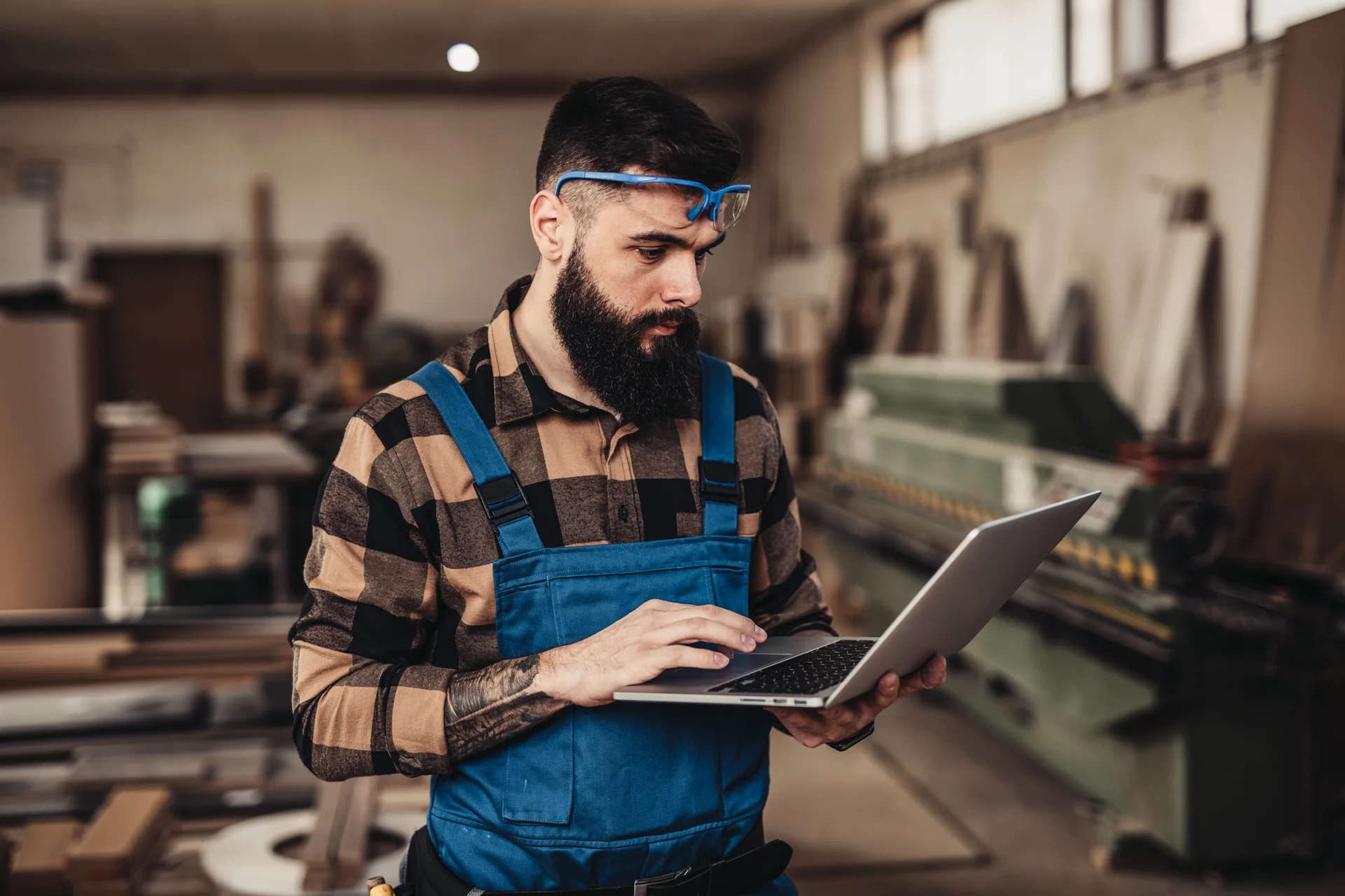 Ein Mann mit einem Bart und blauen Arbeitsoverall, der in einer Werkstatt an einem Laptop arbeitet. Er schaut aufmerksam auf den Bildschirm und ist von Maschinen und Materialien umgeben.