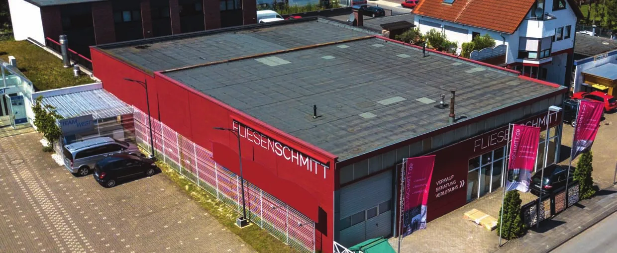 Das Bild zeigt ein großes Gebäude mit roter Fassade, auf dessen Vorderseite gut sichtbar der Schriftzug „FLIESENSCHMITT“ angebracht ist. Es handelt sich offenbar um ein Geschäftsgebäude, möglicherweise ein Laden oder eine Ausstellungshalle. Vor dem Gebäude sind mehrere Autos geparkt, und der Bereich ist mit Pflastersteinen befestigt. Im Hintergrund befinden sich weitere Gebäude, darunter ein Wohnhaus mit schrägem Dach. Rund um den Eingang sind zudem Banner und Grünflächen zu sehen. Insgesamt wirkt die Umgebung wie ein Gewerbegebiet.