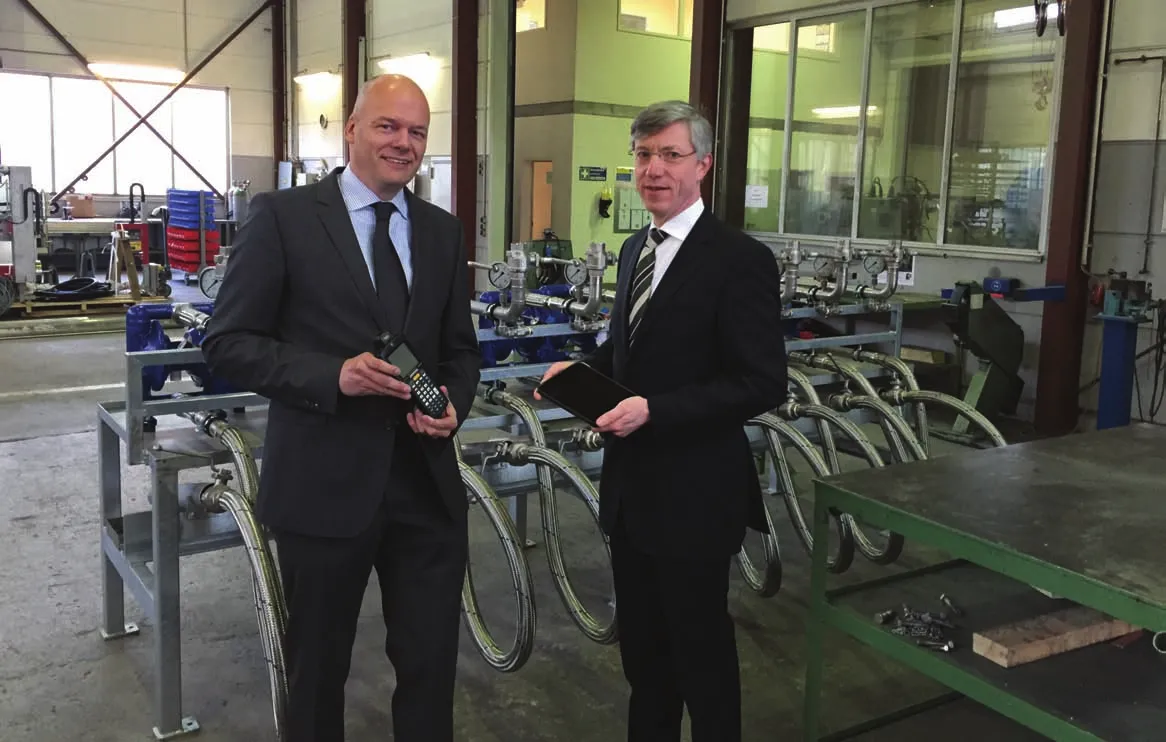 Das Bild zeigt zwei Männer in formeller Kleidung, die sich in einer Werkstatt oder industriellen Umgebung befinden. Einer der Männer hält ein Gerät in der Hand, möglicherweise einen Handscanner oder ein mobiles Computergerät, während der andere ein Tablet hält. Im Hintergrund sind verschiedene Industrieanlagen und Maschinen zu sehen, darunter Rohrleitungen und Ventile. Die Umgebung ist gut beleuchtet und ordentlich, typisch für eine Fertigungs- oder Ingenieurseinrichtung.