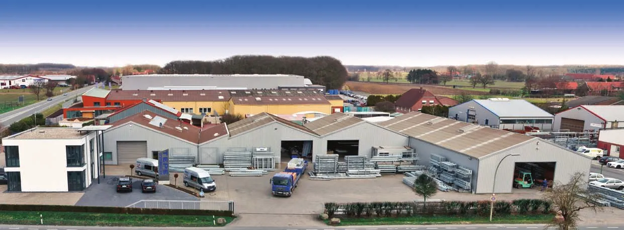 Das Bild zeigt ein Industriegebiet mit mehreren Gebäuden, darunter Lagerhallen und Bürogebäude. Vor den Gebäuden sind Fahrzeuge geparkt, und im Außenbereich sind verschiedene Materialien oder Ausrüstungen zu sehen. Die Umgebung deutet auf einen Geschäfts- oder Produktionsstandort hin, im Hintergrund erstrecken sich Felder und Bäume. Der Himmel ist klar, was auf einen sonnigen Tag hinweist.