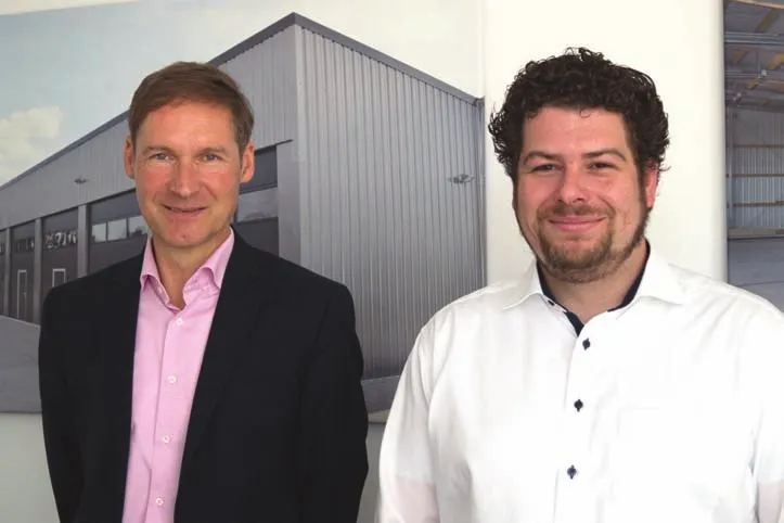 Zwei Männer, die nebeneinanderstehen und lächeln. Einer trägt einen dunklen Anzug mit einem hellrosa Hemd, der andere ein weißes Hemd. Im Hintergrund ist ein großes Bild eines Gebäudes oder einer Struktur zu sehen, vermutlich ein Lagerhaus oder eine Industrieanlage. Die Umgebung wirkt wie ein Büro oder eine professionelle Arbeitsumgebung.