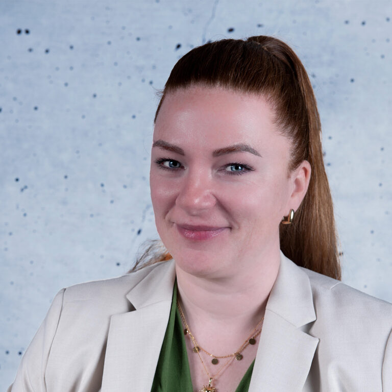 Ein Porträt einer Frau mit langen, roten Haaren, die zu einem Pferdeschwanz gebunden sind. Sie trägt ein helles Jackett über einem grünen Oberteil und sieht mit einem freundlichen Lächeln in die Kamera. Der Hintergrund besteht aus einem blauen, unscharfen Muster.